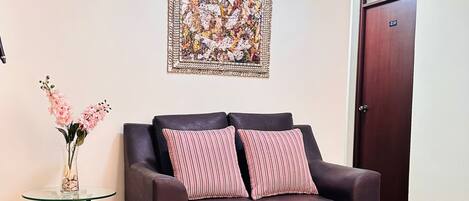 Un sofá de cuero marrón oscuro con dos almohadones a rayas, una mesa de vidrio con un jarrón de flores rosas y un cuadro abstracto grande en la pared.