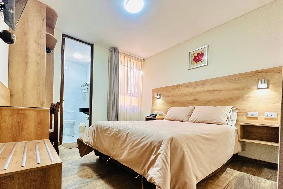 A hotel room with a wooden headboard, a bed with beige bedding, a bedside table, and a small nightstand.