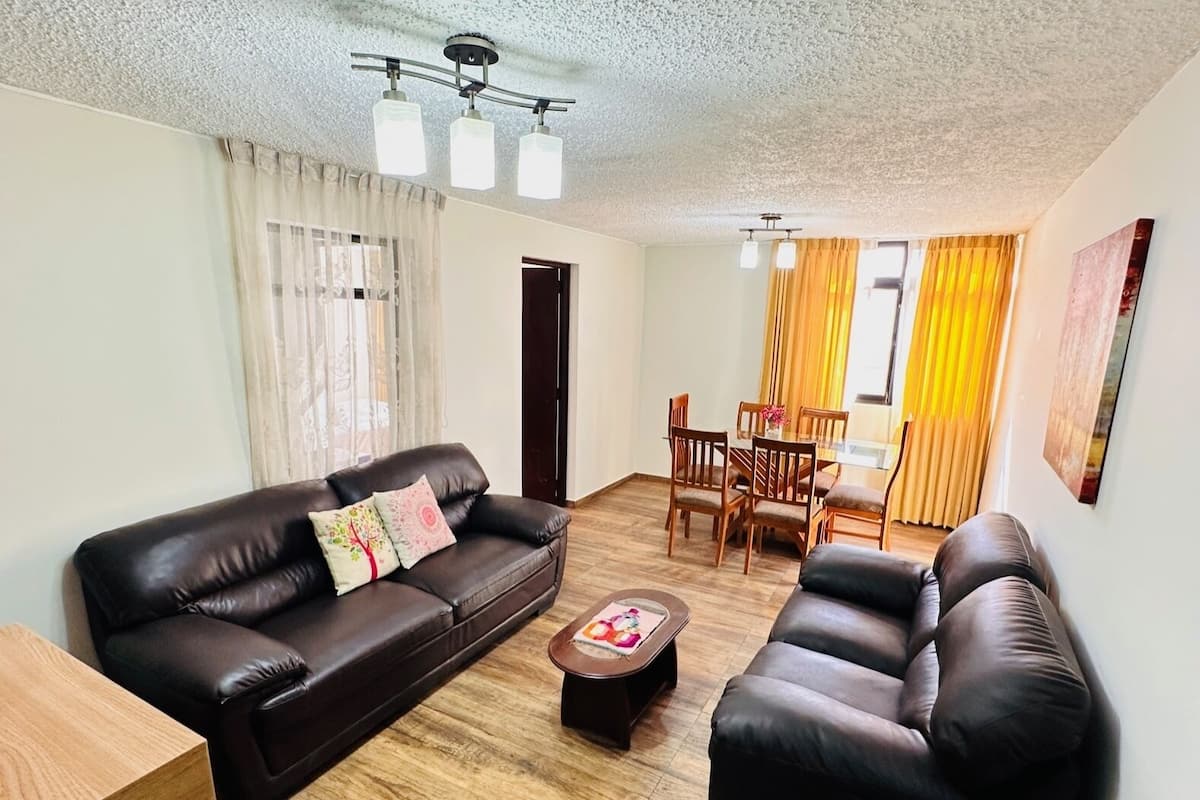 A living room with a brown leather sofa, a wooden coffee table, and a dining area with yellow curtains and wooden chairs.