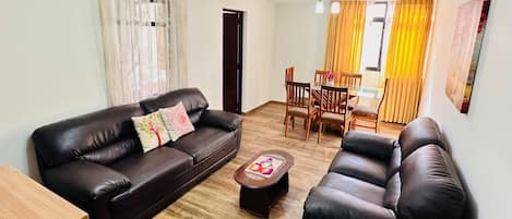 A living room with a brown leather sofa, a wooden coffee table, and a dining area with yellow curtains and wooden chairs.