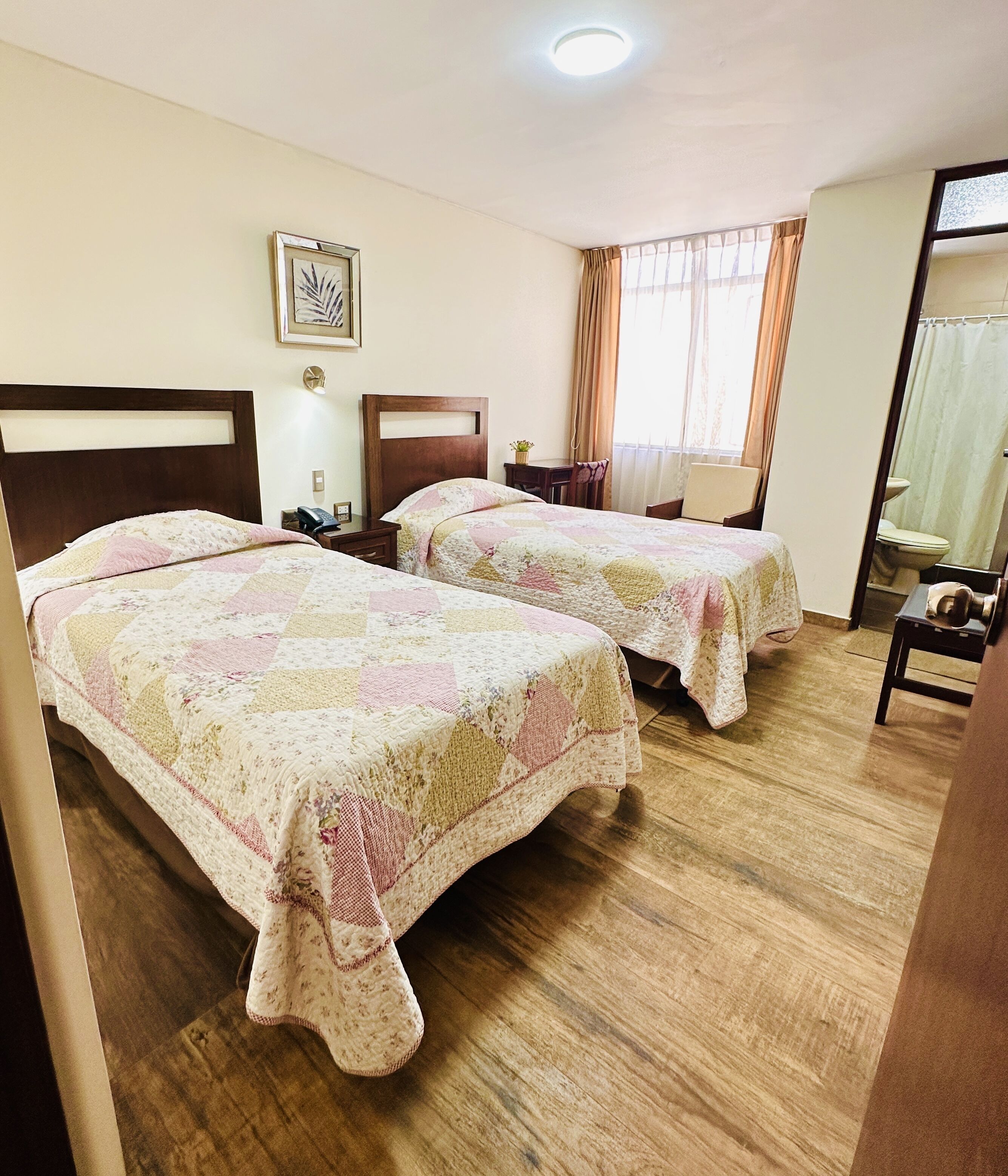 A hotel room with two beds, wooden flooring, a small table, a chair, a nightstand, a lamp, a framed picture on the wall, and a window with curtains.