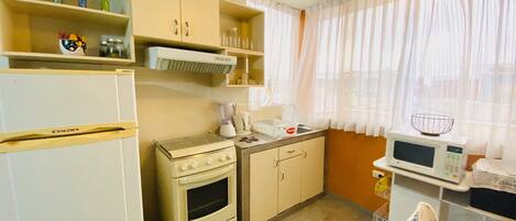 A compact kitchen with a refrigerator, stove, microwave, and a window with sheer curtains.