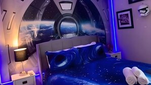 Une chambre avec une fenêtre ronde, un lit orné de motifs étoilés, une table de chevet avec une lampe et un mur décoré de tableaux encadrés.