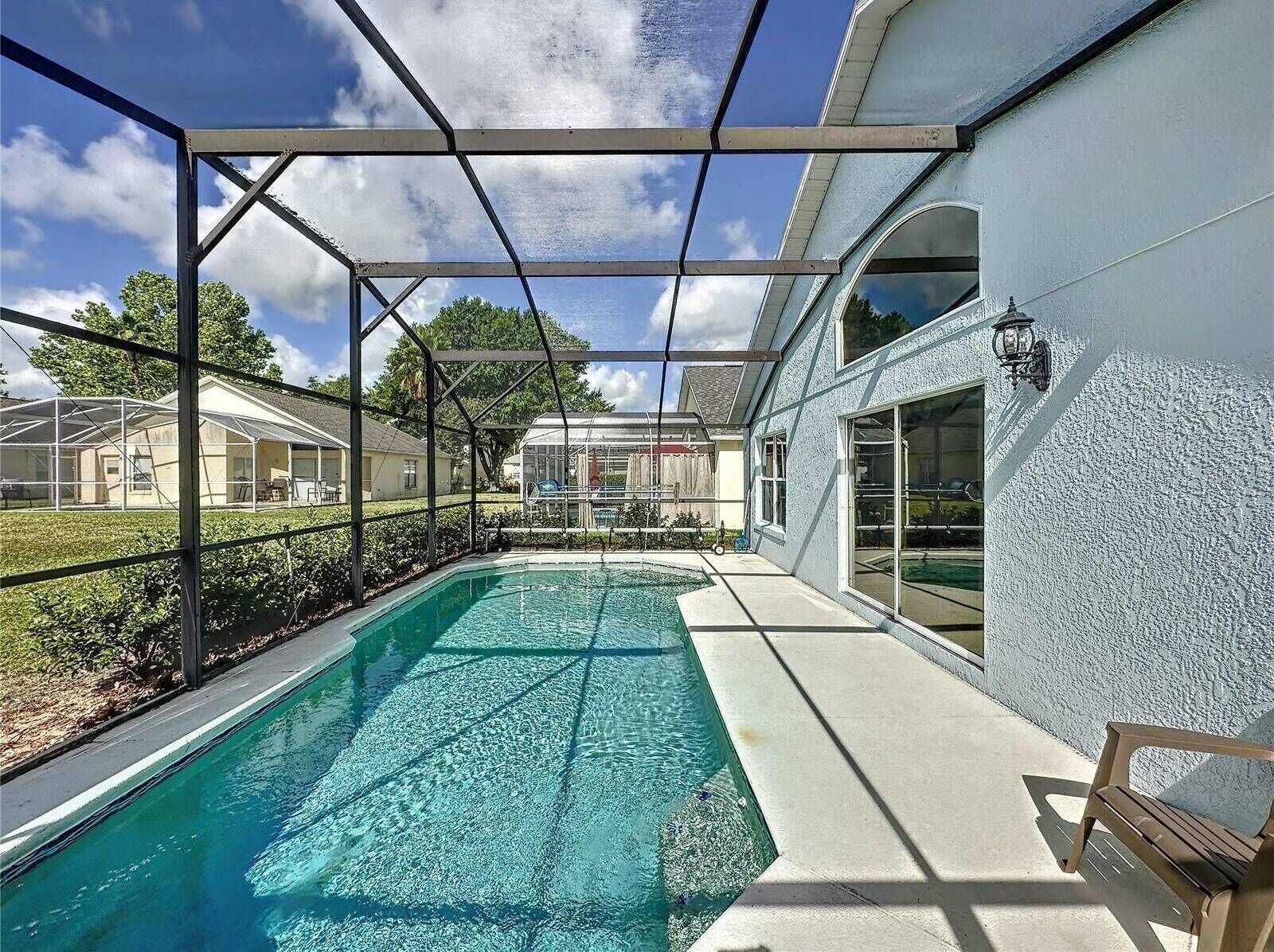 A swimming pool with a covered area, surrounded by a fence, and adjacent to a building with large windows and a light fixture.