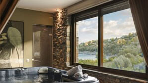 A spa room with a hot tub, stone wall, and a window overlooking a scenic landscape. - Paramithenio Village Beach Resort & Spa (Kymi-Aliveri)