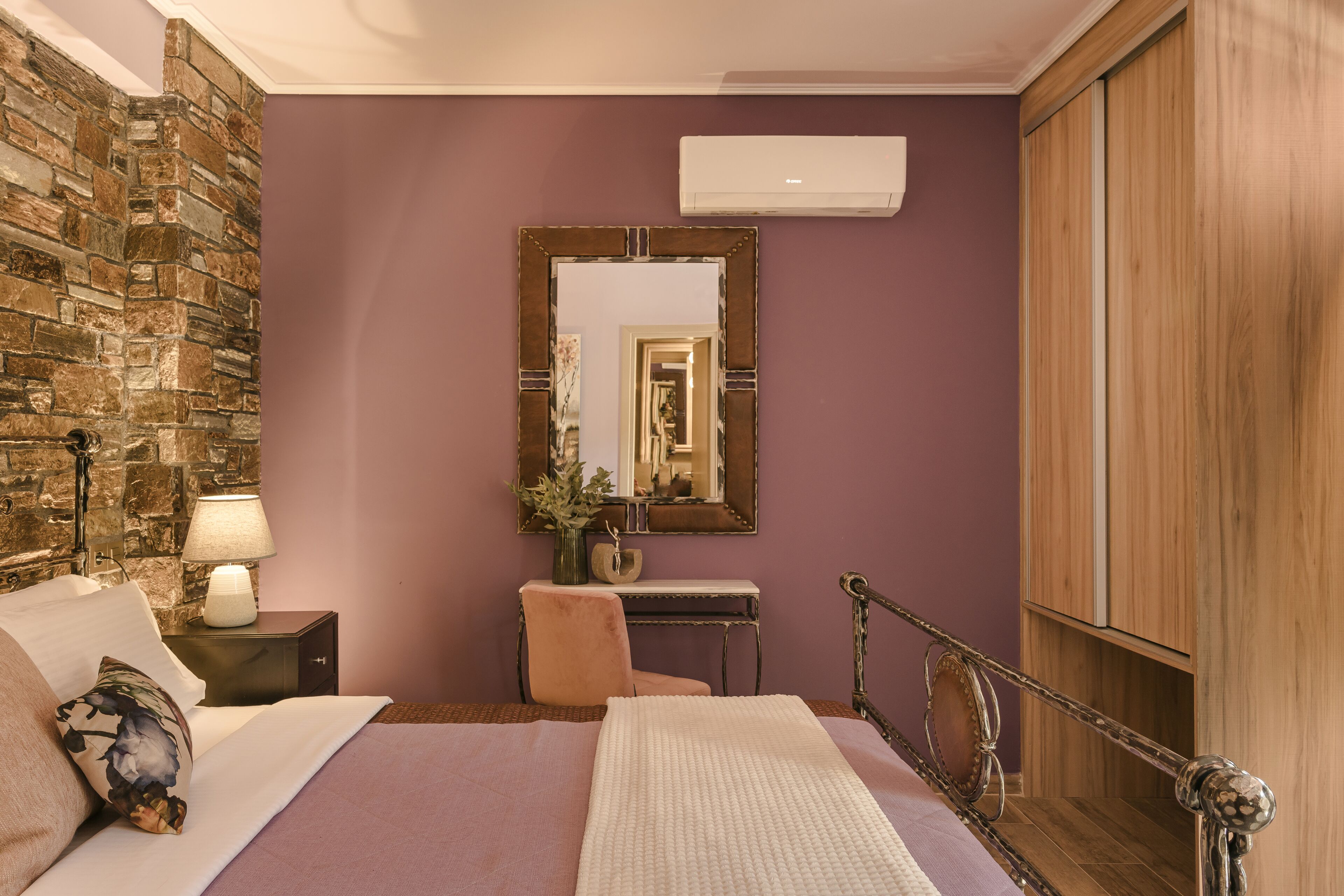 A bedroom with a stone wall, a bed with a white comforter, a nightstand with a lamp, a mirror, and a wooden wardrobe.