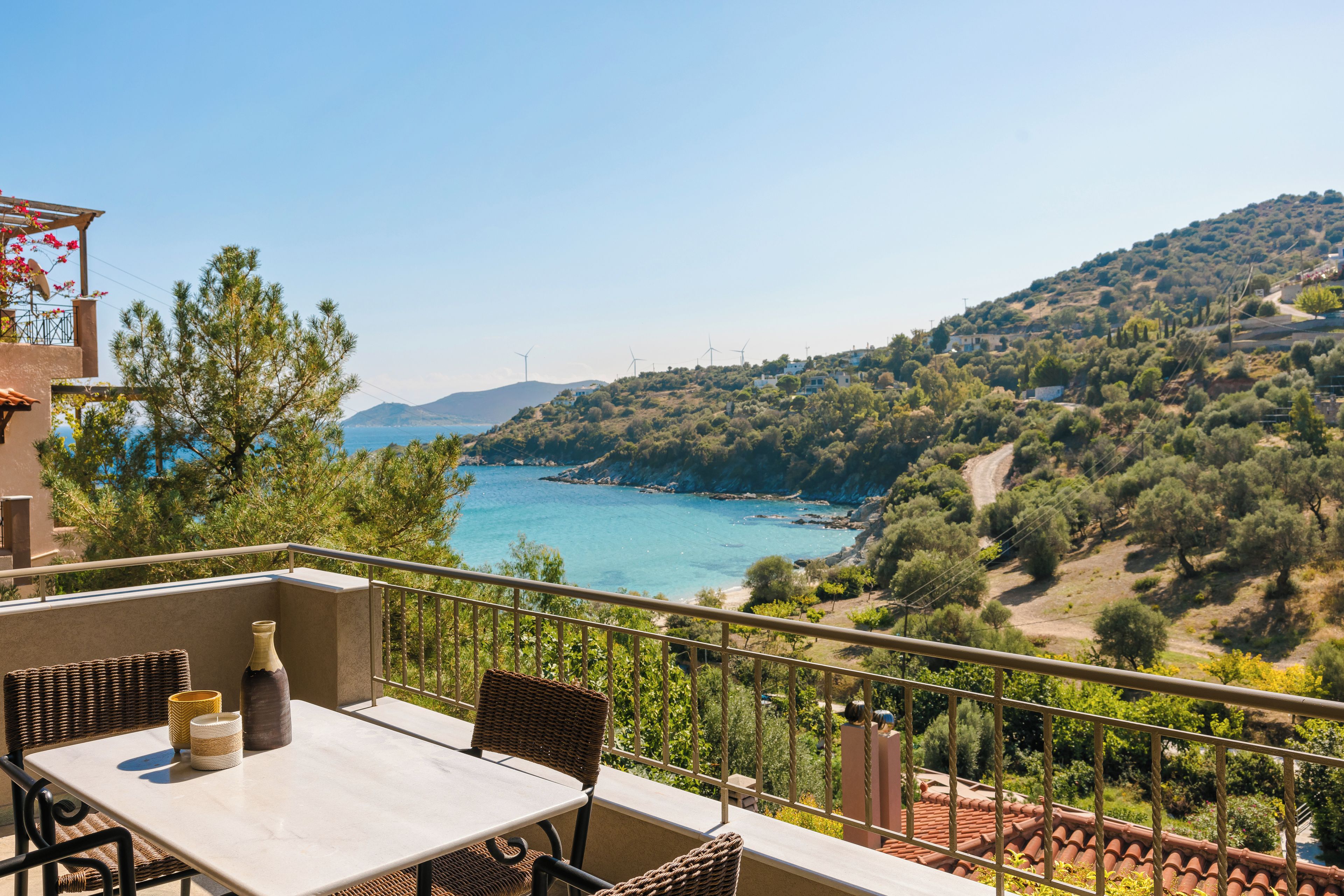 A balcony with a table and chairs overlooking a coastal landscape with wind turbines and a hilly terrain.