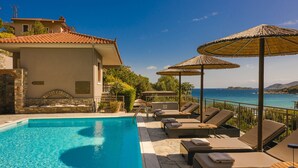 A poolside area with lounge chairs and umbrellas, overlooking a body of water. - Paramithenio Village Beach Resort & Spa (Kymi-Aliveri)