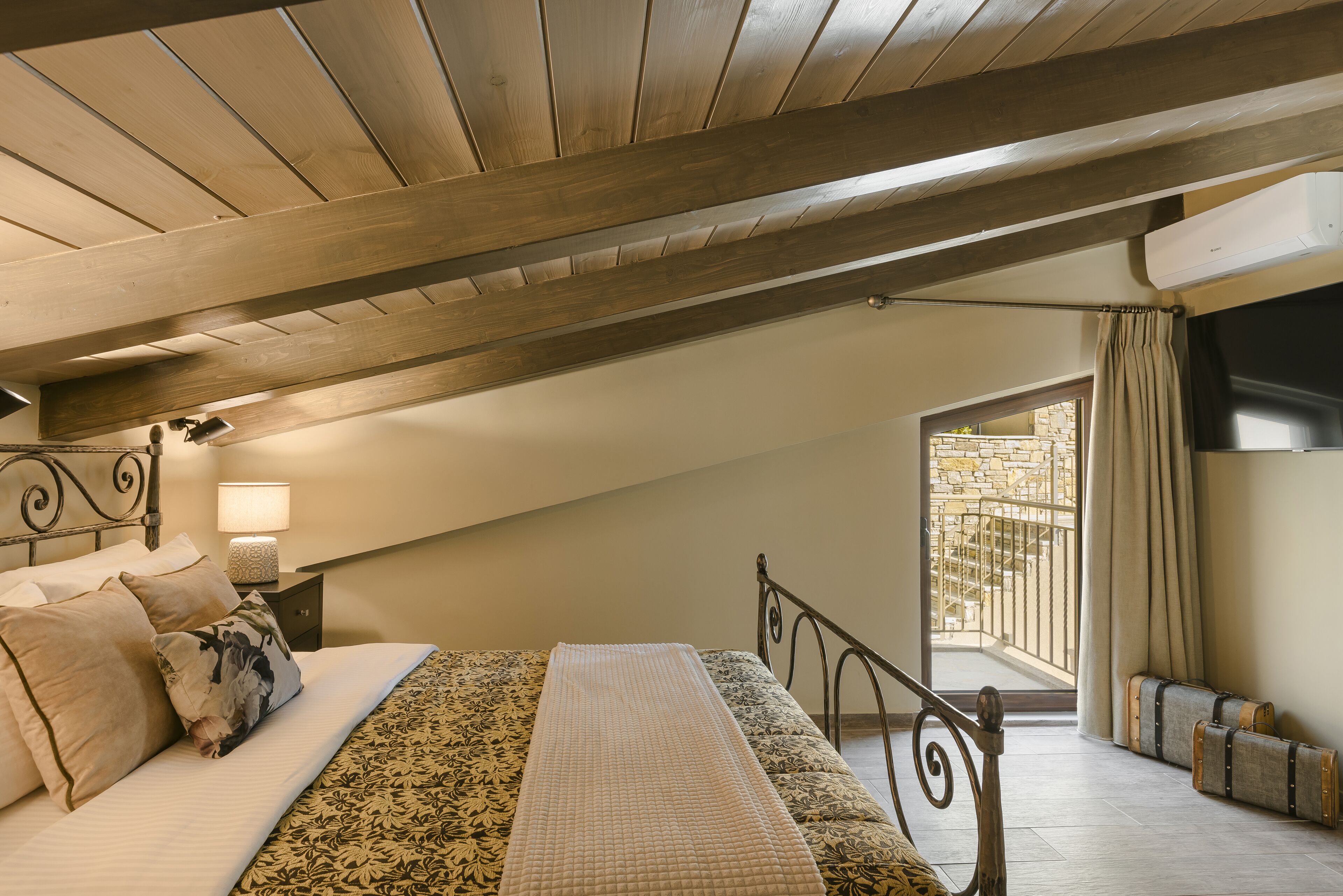 A bedroom with a sloped ceiling, a bed with patterned bedding, a nightstand with a lamp, and a view of a balcony with a stone wall.