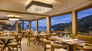 A restaurant with multiple tables and chairs, large windows with a view of the sea and hills, and modern chandeliers. - Paramithenio Village Beach Resort & Spa (Kymi-Aliveri)