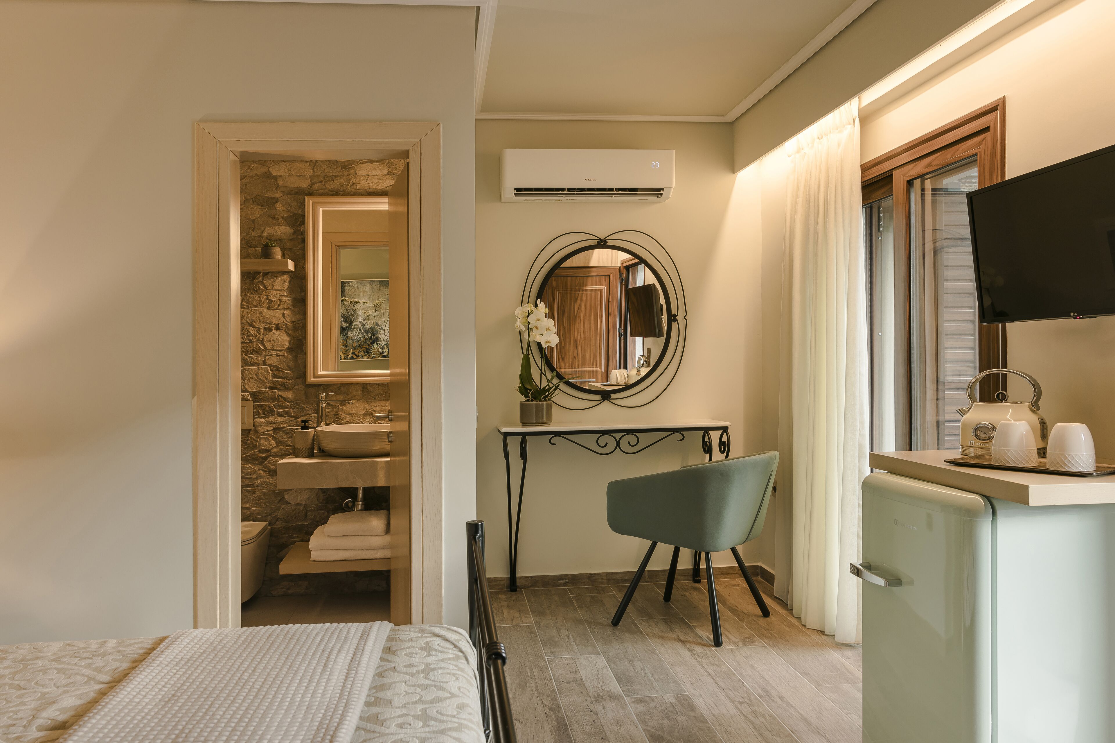 A compact hotel room with a bed, a small desk with a chair, a kitchenette, a bathroom with a stone wall, and a TV.