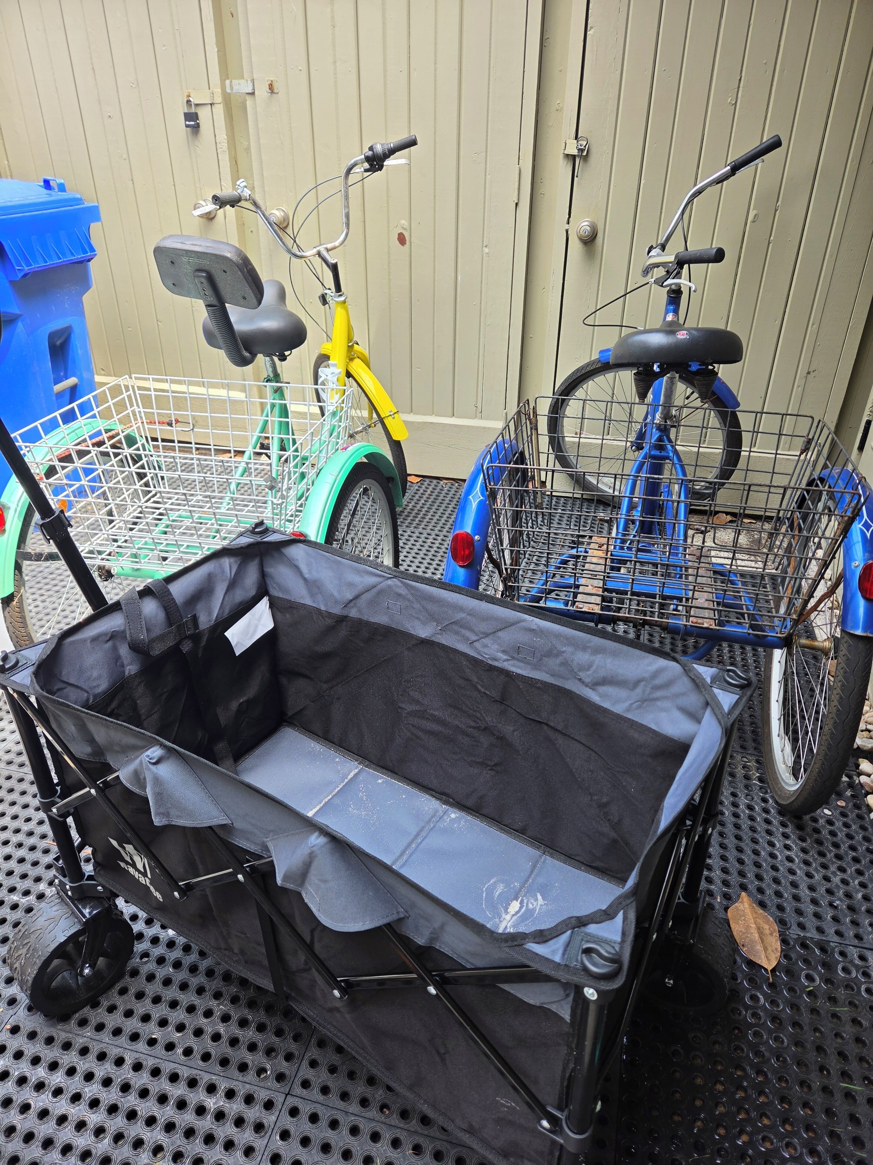 A black wheeled suitcase with a mesh top, a blue bicycle with a basket, and a yellow tricycle with a basket.