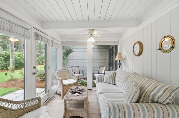A room with a white ceiling, a sofa, a wicker chair, a table with a book and stones, and a view of a garden through a glass door.