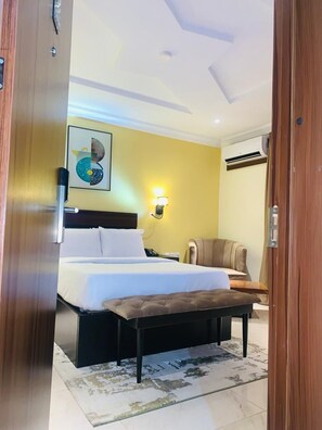 A hotel room with a bed, a bedside table, a chair, a wall-mounted lamp, and a painting.