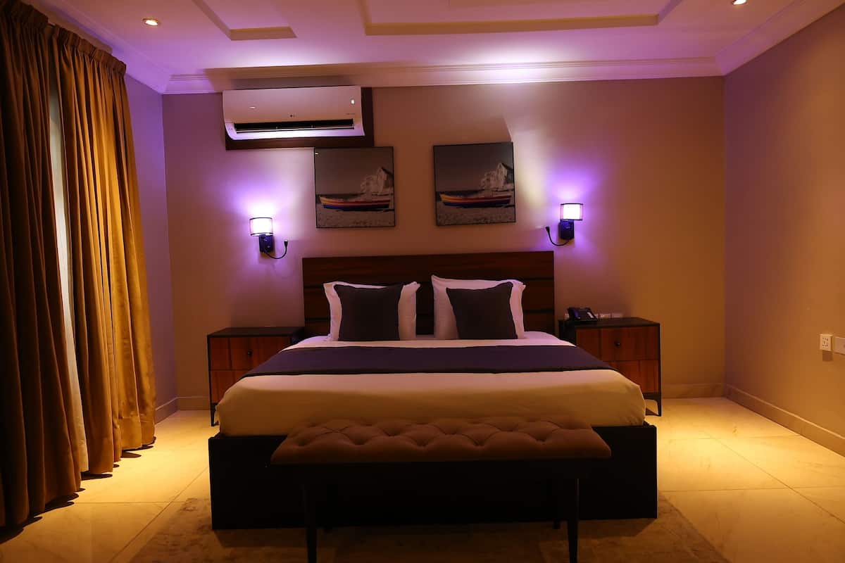 A bedroom with a large bed, two bedside tables, an air conditioner, wall-mounted lights, and a window with curtains.