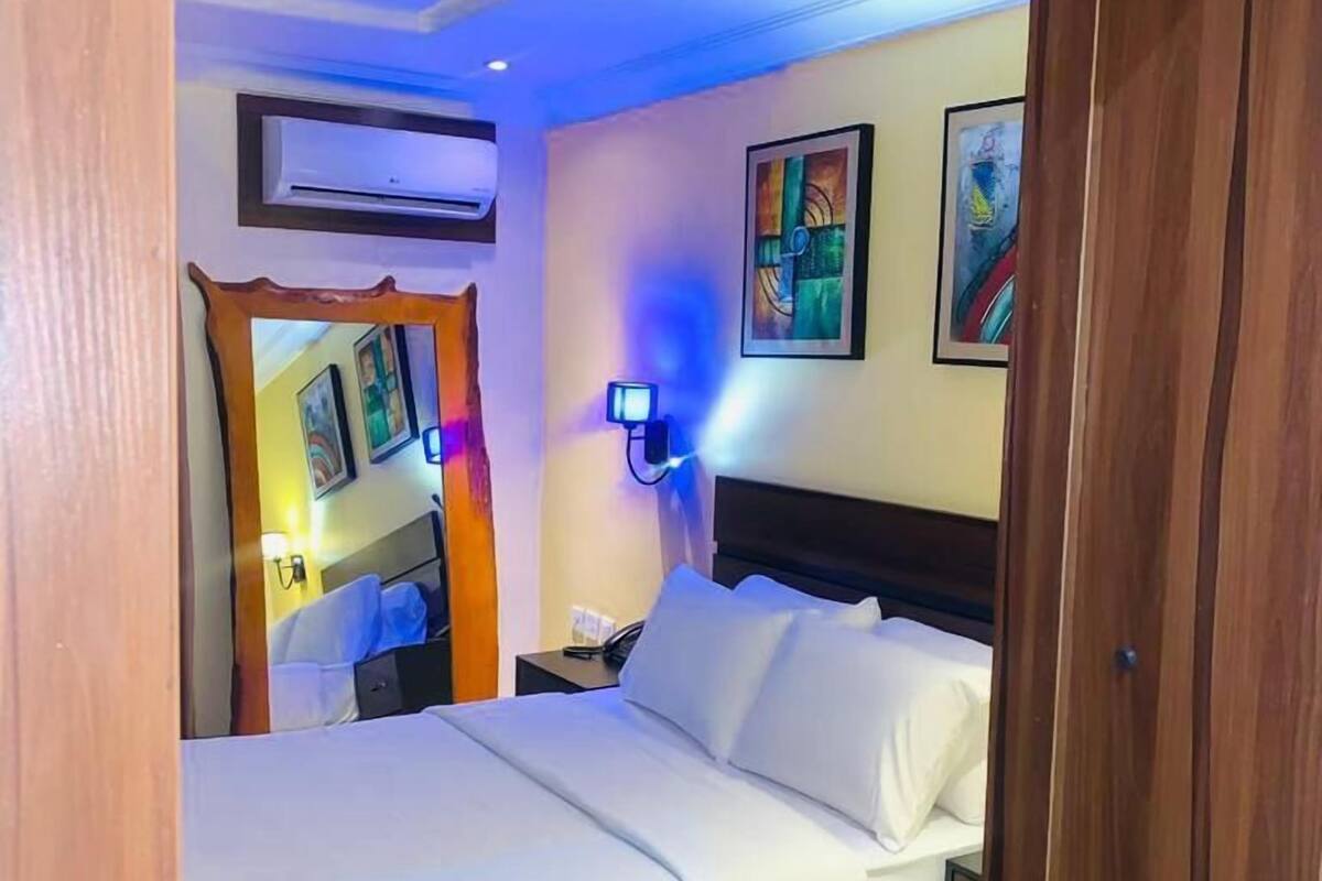 A hotel room with a bed, a nightstand, an air conditioning unit, and artwork on the walls.