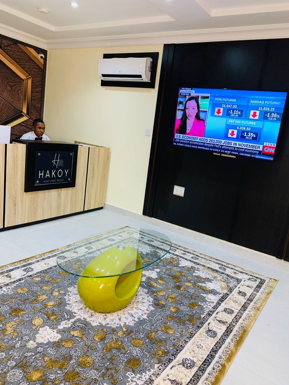 Zona de recepción de hotel con un mostrador identificado como 'HAKOY BOUTIQUE HOTEL', un televisor mostrando noticias financieras y una escultura de vidrio decorativa sobre una alfombra estampada.