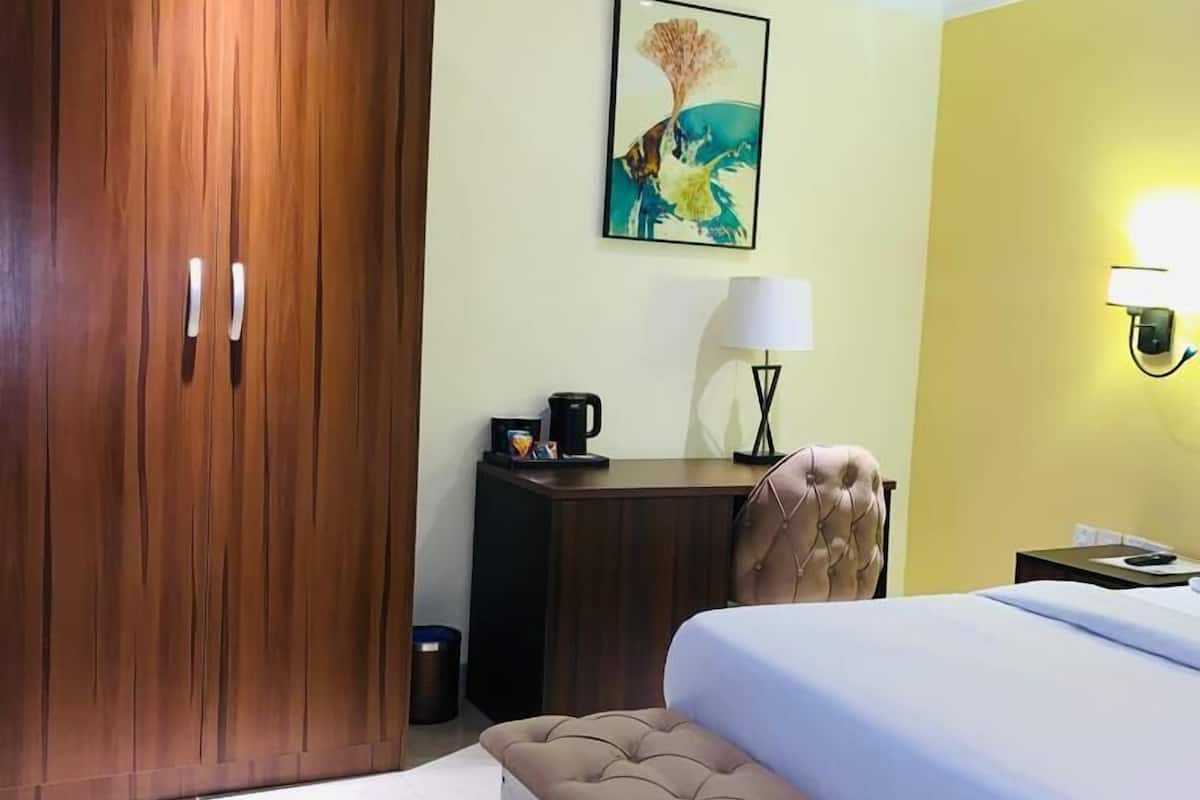 A hotel room with a large bed, a wooden wardrobe, a bench, a desk with a lamp, and a painting on the wall.