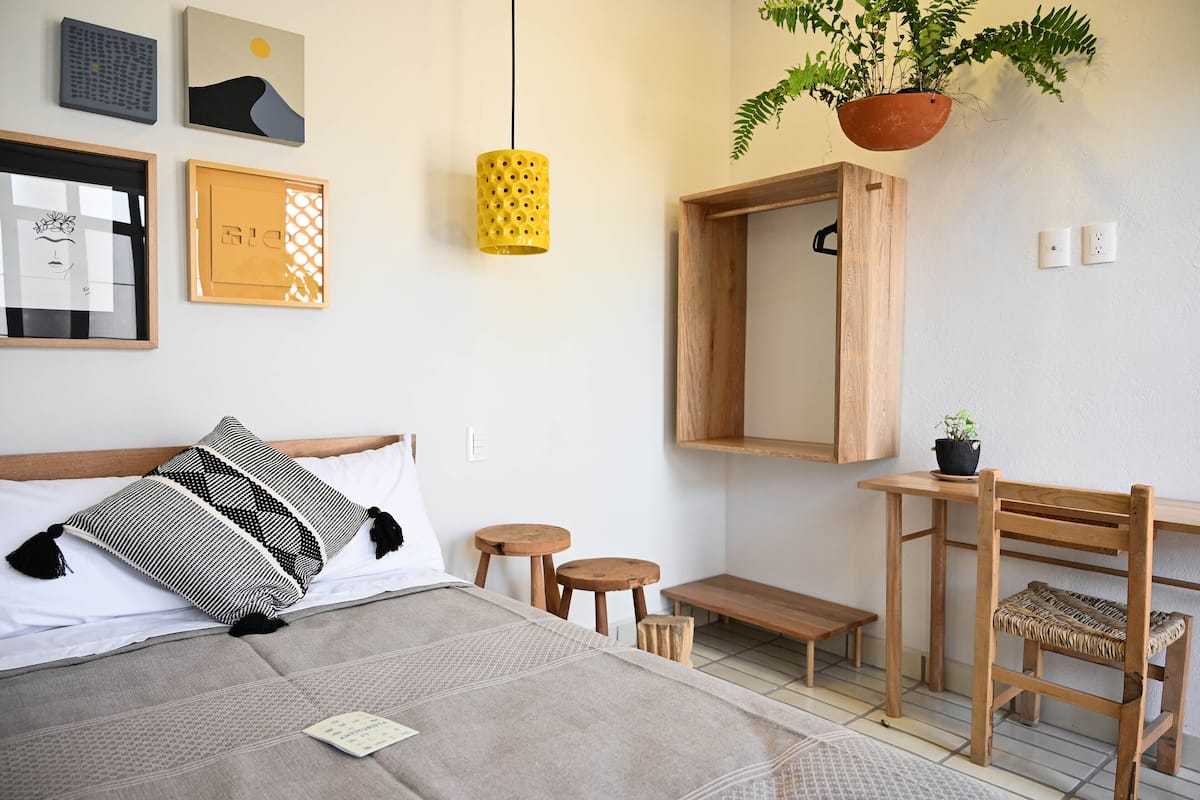 Une chambre à coucher bien rangée, avec un lit, des tables de chevet, une chaise en bois, une plante suspendue et des œuvres d’art murales.