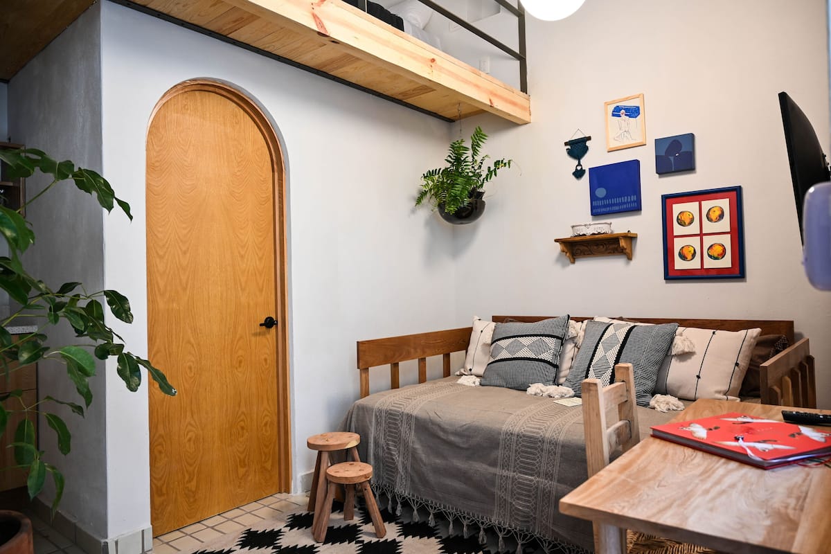 Une chambre confortable avec une porte en bois, un lit avec une literie à motifs, un petit tabouret, une table avec un dossier rouge et une plante suspendue.