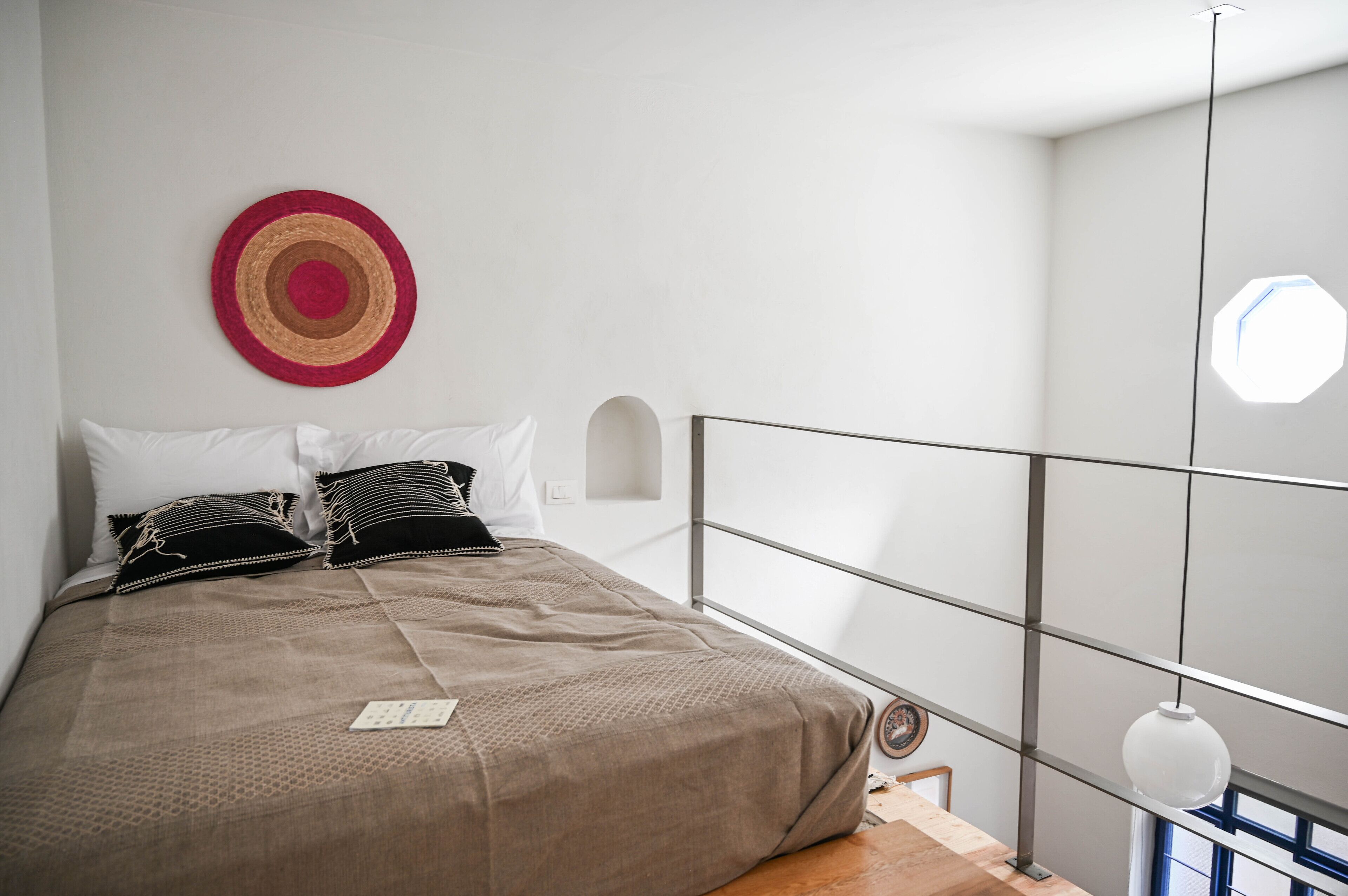 Een net opgemaakt bed met een bruine deken en twee kussens, een ronde wanddecoratie, en een raam met uitzicht op de lucht.