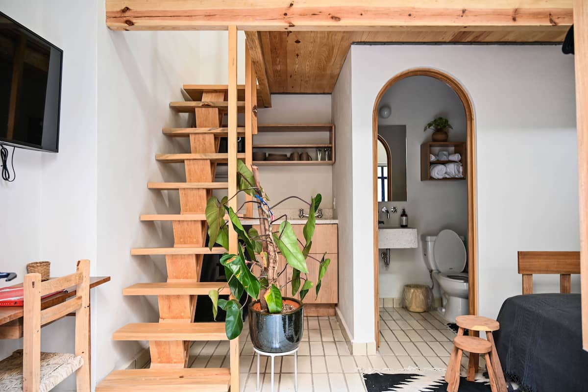Une pièce de taille modeste, avec un escalier en bois, une plante en pot, un lit et un petit tabouret.