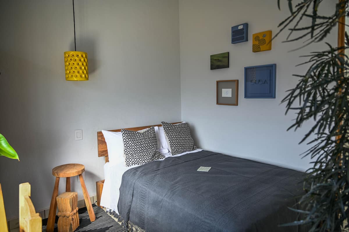 Une chambre à coucher bien rangée, avec un lit, un tabouret en bois, une lampe suspendue et des tableaux accrochés au mur.