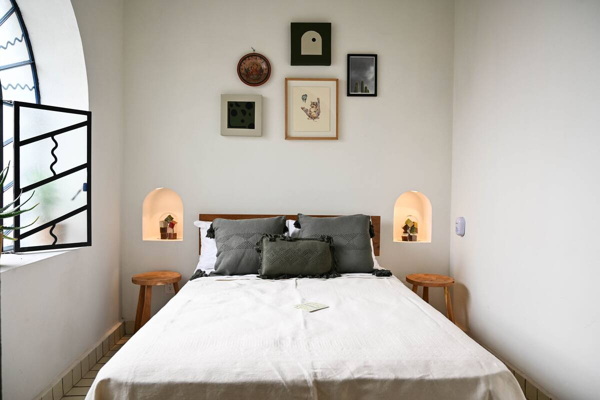 Une chambre à coucher bien rangée, avec un lit, deux tables de chevet et une étagère murale agrémentée d’objets décoratifs.