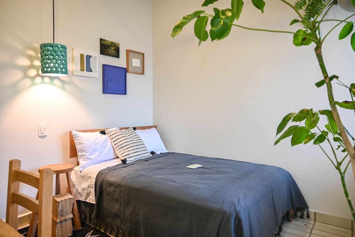 Un lit bien fait, recouvert d’une couverture bleue, une chaise en bois, une lampe verte suspendue et un mur orné de tableaux encadrés.