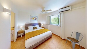 A neatly arranged hotel room with a bed, bedside lamps, a ceiling fan, an air conditioning unit, a window with curtains, and a chair.