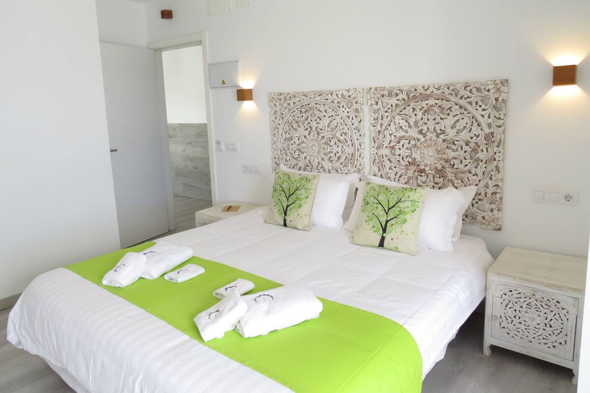 A neatly made bed with green bedspread and white pillows, a wooden nightstand with intricate carvings, and a wall-mounted light fixture.