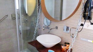 A bathroom with a glass shower enclosure, a white sink, a wooden shelf with a blue leaf decoration, and a round mirror with a clock. - Hotel Lua Isla Del Hierro (El Pinar de El Hierro)