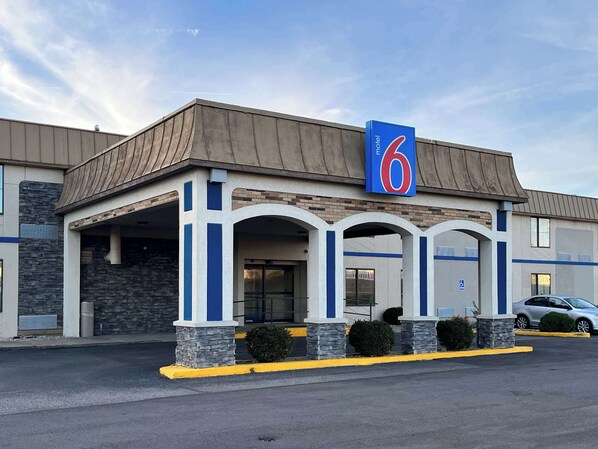 A motel with a '6' sign, a covered entrance, stone pillars, and a parking lot with cars.