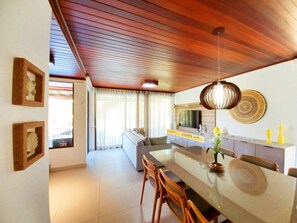 A modern living room with a wooden ceiling, a dining area with a long table, and a kitchen with a TV.