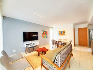 A modern living room with a flat-screen TV, a sofa, a dining table with chairs, and two abstract paintings on the wall. - Lindos Bangalôs no Eco Resort  (Tamandaré)