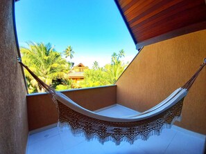 A hammock on a balcony with a view of palm trees and a building. - Lindos Bangalôs no Eco Resort  (Tamandaré)