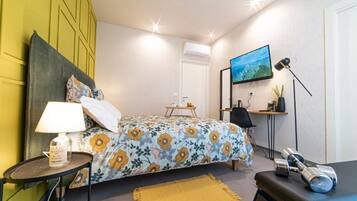 A bedroom with a bed, a bedside table with a lamp, a TV on the wall, and a small desk with a chair.