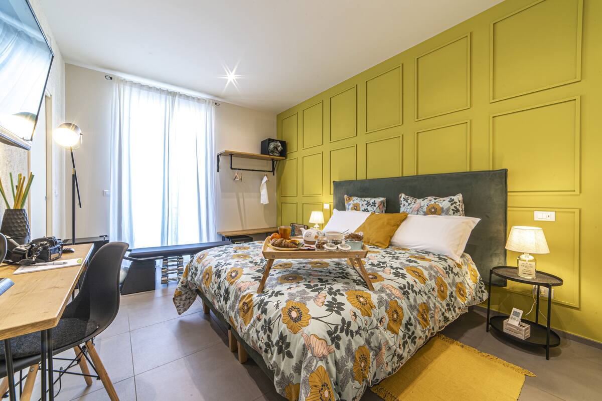 A bedroom with a floral bedspread, a bedside table with a lamp, and a desk with a chair.