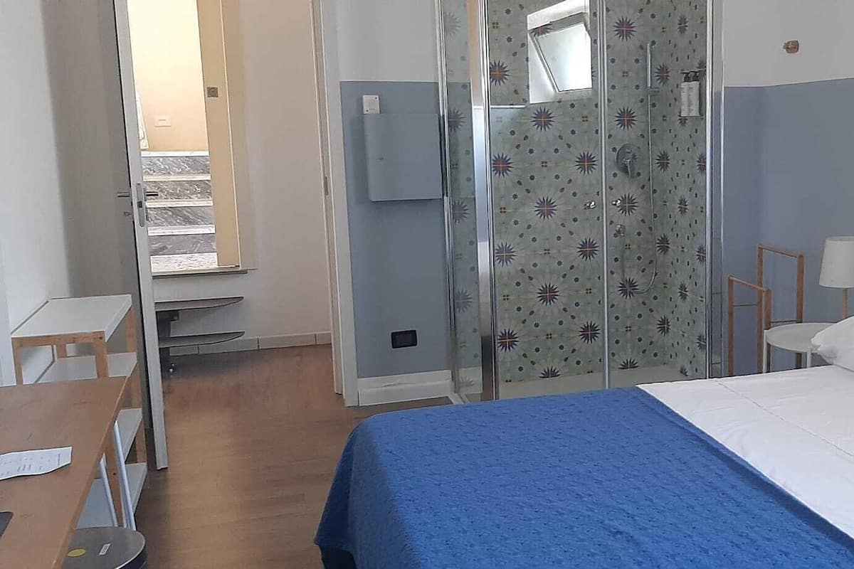 A hotel room with a bed, a desk, a chair, a lamp, and a shower area.