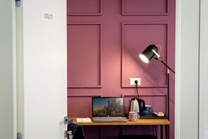 A room with a purple wall, a desk with a laptop, a coffee maker, and a lamp.