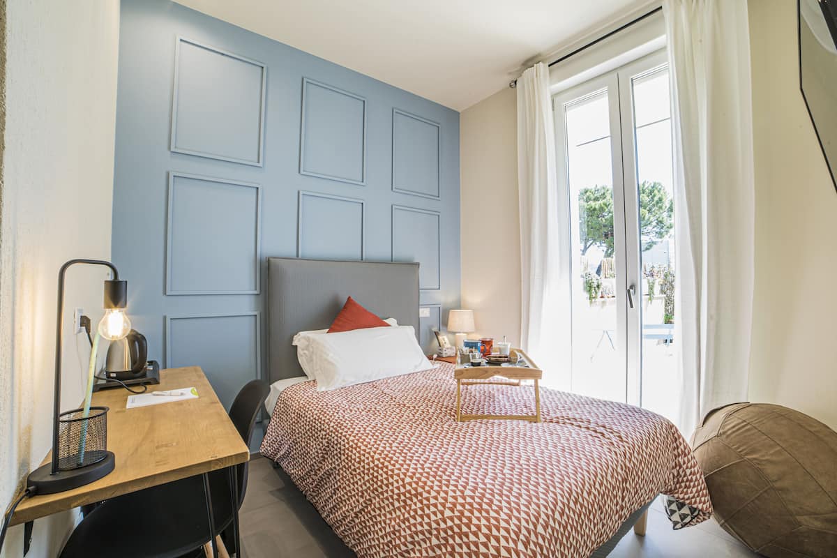 A bedroom with a bed, a desk with a lamp, a nightstand, and a window with curtains.