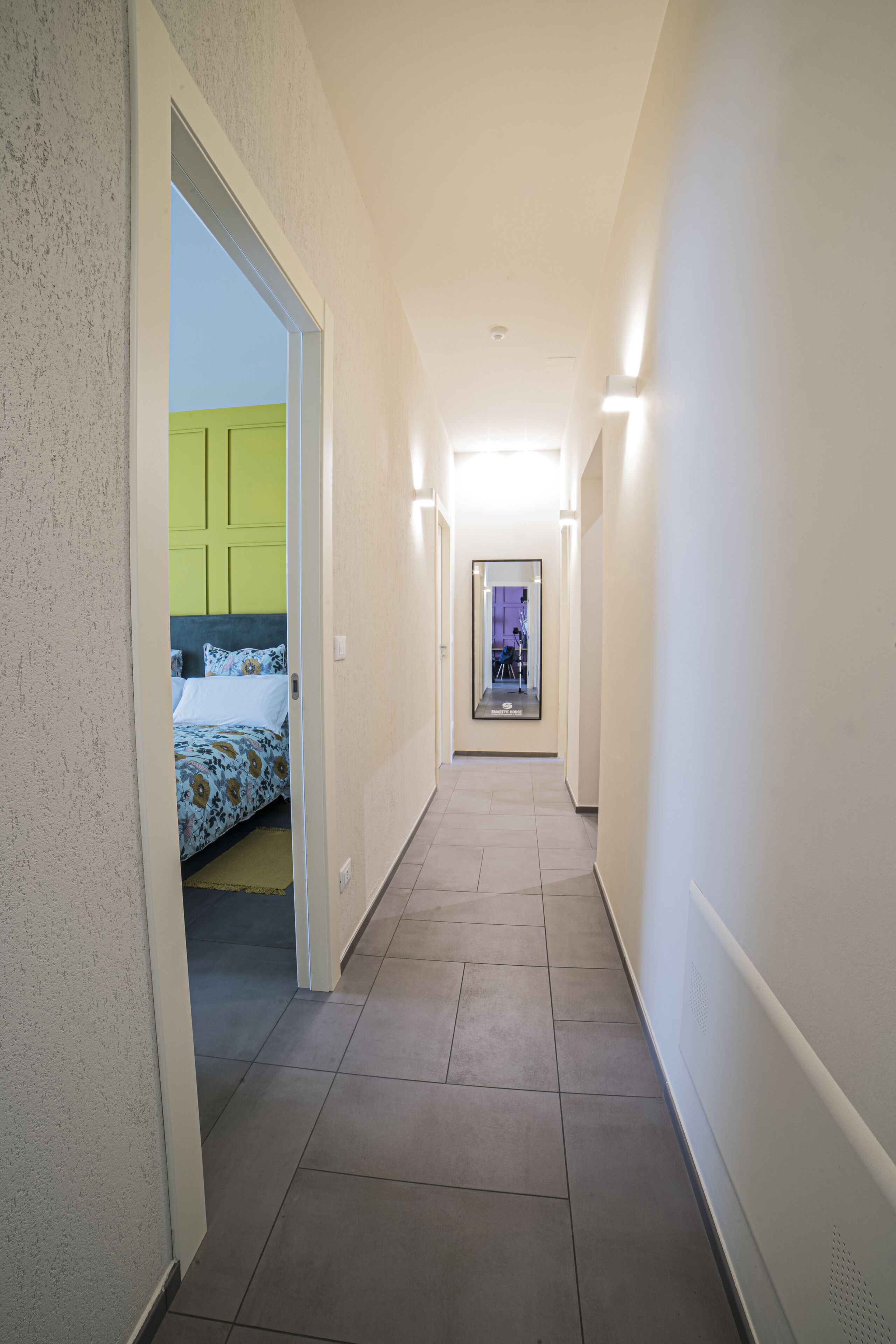 A narrow hallway with a bed at the end, a door with a green panel, and a mirror reflecting a room with a desk and chair.