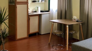 Eine kleine Küche mit Kühlschrank, Spüle und einem kleinen Esstisch mit Hocker. Eine Topfpflanze und ein Bild schmücken die Wand.