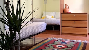 Ein Schlafzimmer mit einem Bett, einer Kommode, einer Topfpflanze, einem farbenfrohen Teppich und einer Wanduhr.
