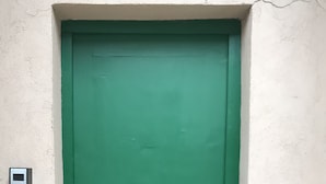 Eine grüne Tür mit Metallbeschlag und Schlüsselloch, eingelassen in eine weiße Wand, links daneben eine kleine Metallkassette.