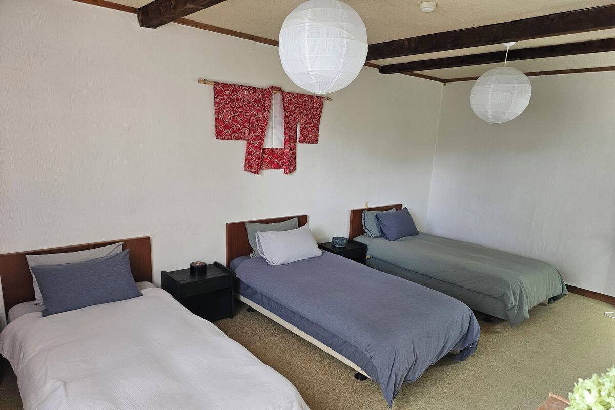 A room with three beds, each with a different colored bedspread. There are two nightstands with lamps and a small table with a bowl. The room has a wooden ceiling with exposed beams and two hanging paper lanterns.