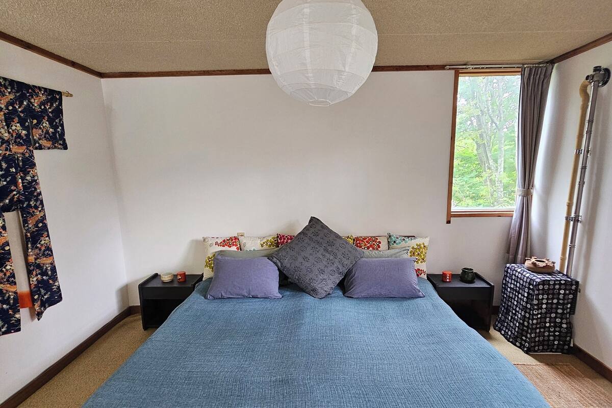 A bedroom with a large bed, a ceiling fan, a window with curtains, and a nightstand with a lamp.