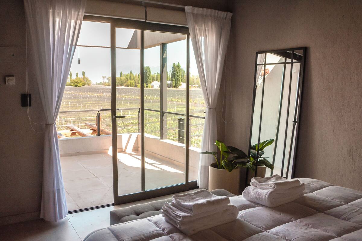 Quarto de hotel com uma grande porta de correr de vidro que dá para uma varanda, uma cama com toalhas bem dobradas, uma planta em vaso e um espelho na parede.