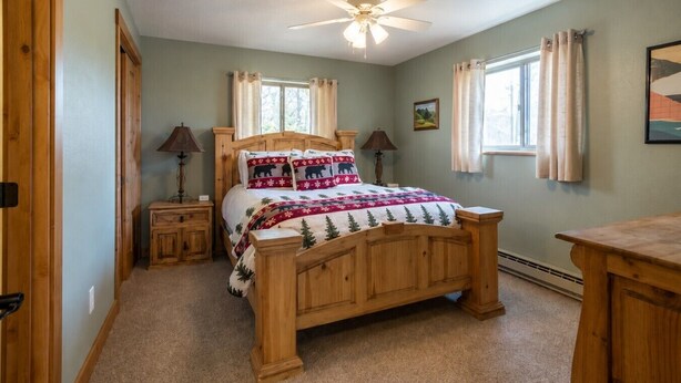 A bedroom with a wooden bed, two bedside tables, a ceiling fan, and windows with curtains. - Mountain Views from WP Top Floor Getaway (Winter Park)