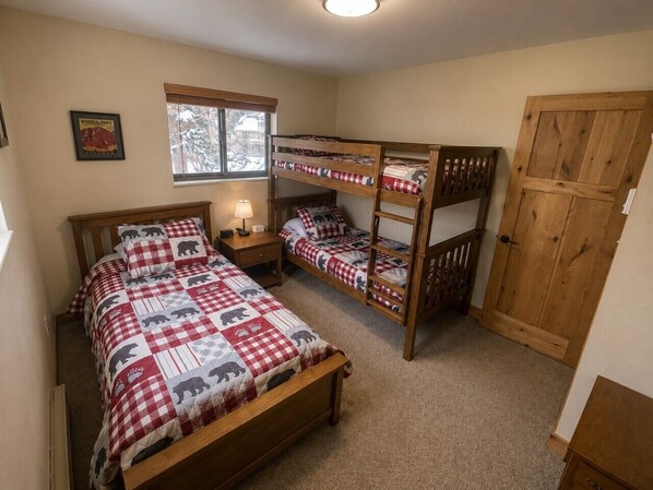 A bedroom with two bunk beds, a single bed, a nightstand with a lamp, a window with blinds, and a door.